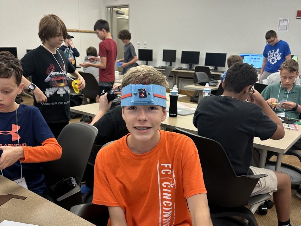 A middle school child wears a handmade microbit-powered headband with fellow campers in the background.