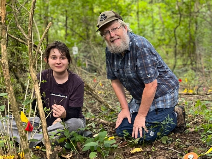 NKU Biological Sciences Undergraduate Research Biology faculty and students sit in a vibrant, green woodland area, measuring honeysuckle growth.