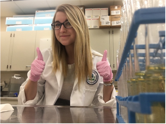 Sarah Goodrich gives two thumbs up in her lab coat and safety gloves.