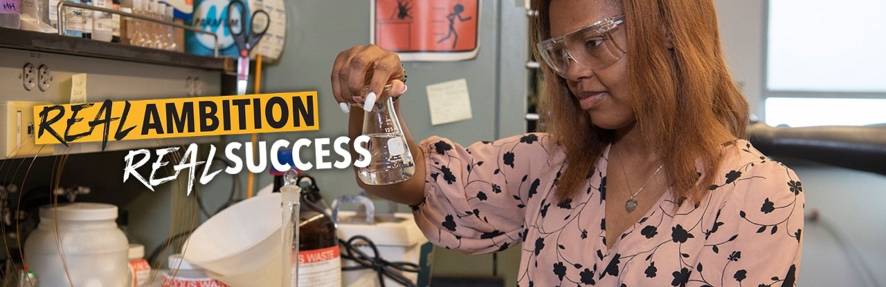 Student wearing protective goggles, observing a beaker inside a chemistry lab.